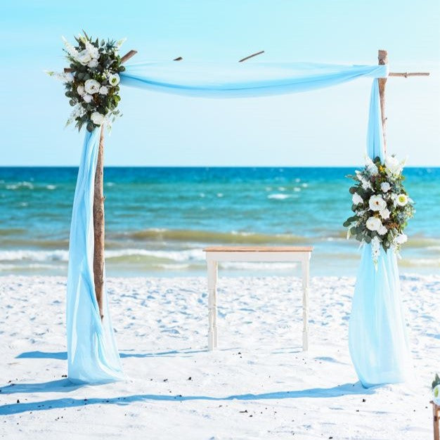 Beach wedding arch with floral decorations and blue drapes against a clear blue sky.