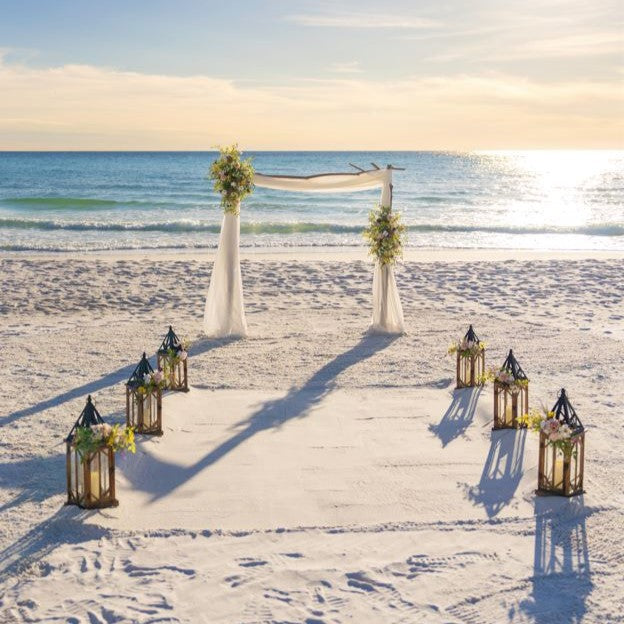 Beach wedding setup with arch, lanterns, and wildflowers on a sandy beach.