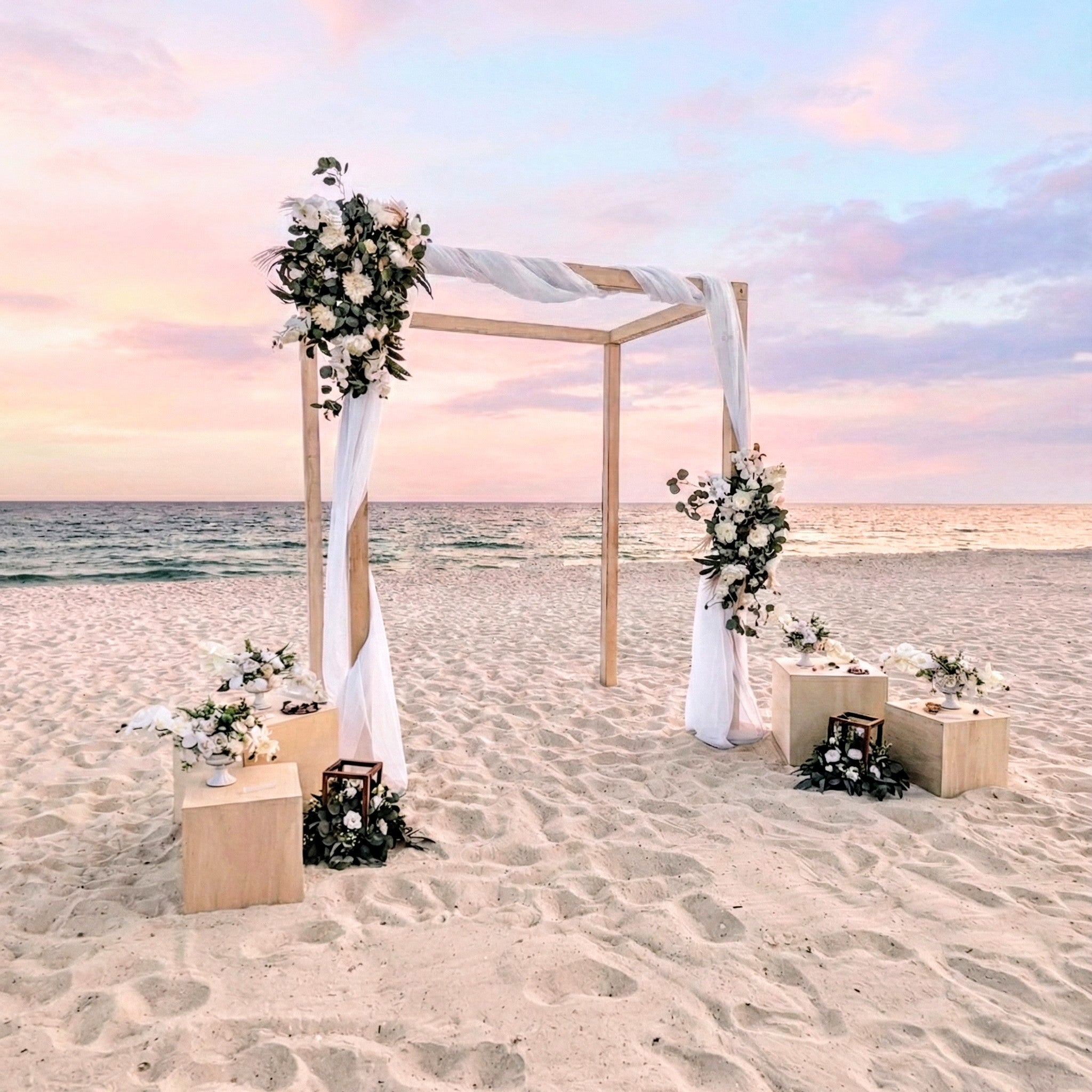 Beach wedding setup with floral arch and decorations on a sandy beach at sunset.