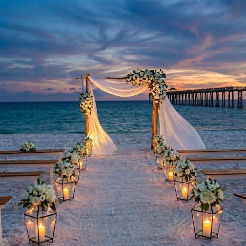 Decorated wedding arch on a beach with lanterns and flowers at sunset.
