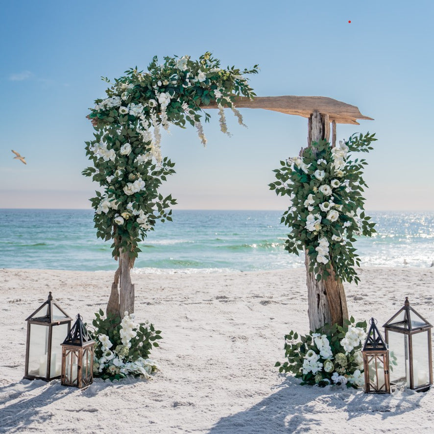 Driftwood wedding arch with flowers and lanterns on a sandy beach with ocean view