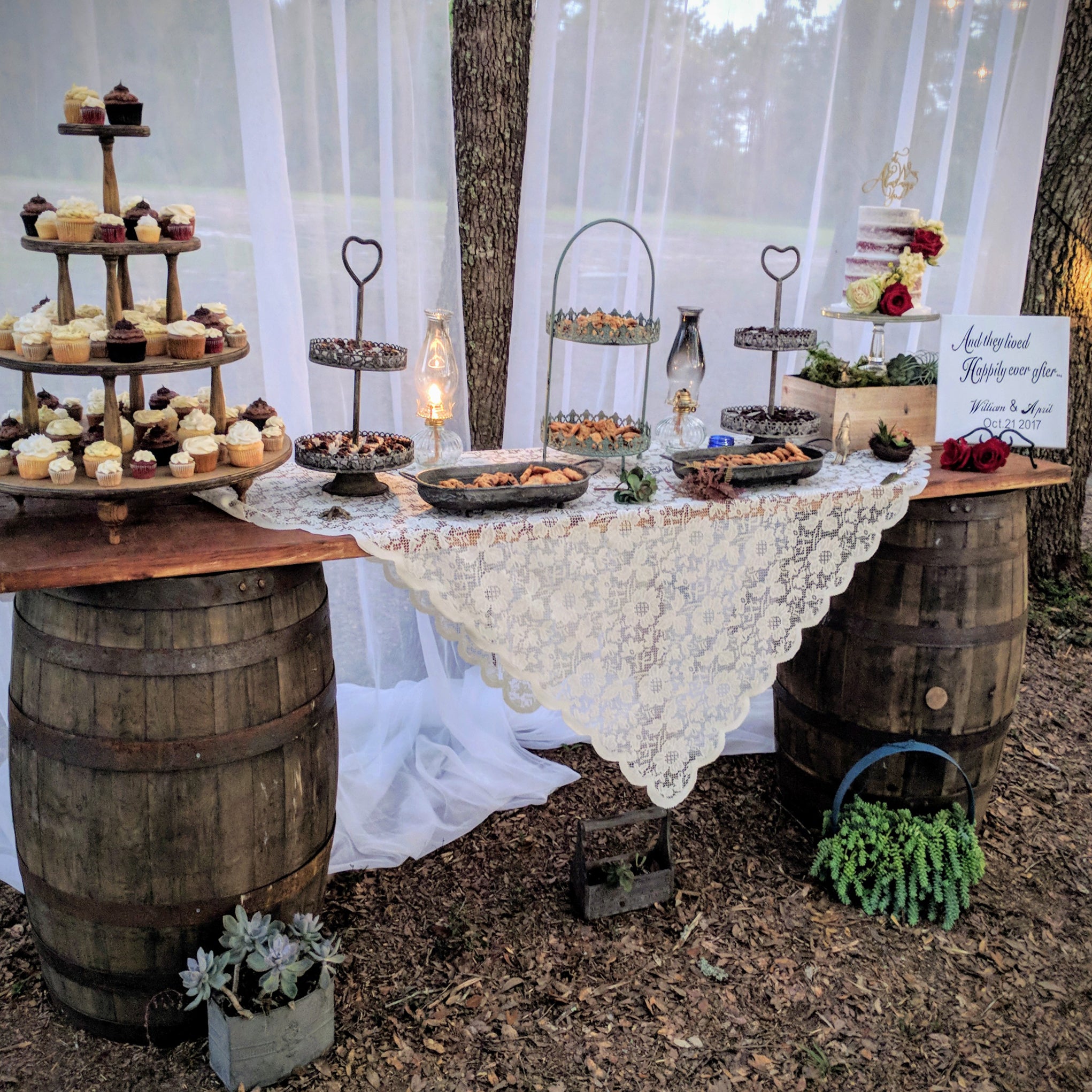 Decorative outdoor table setting with cake stands and a lace tablecloth, surrounded by barrels and plants.