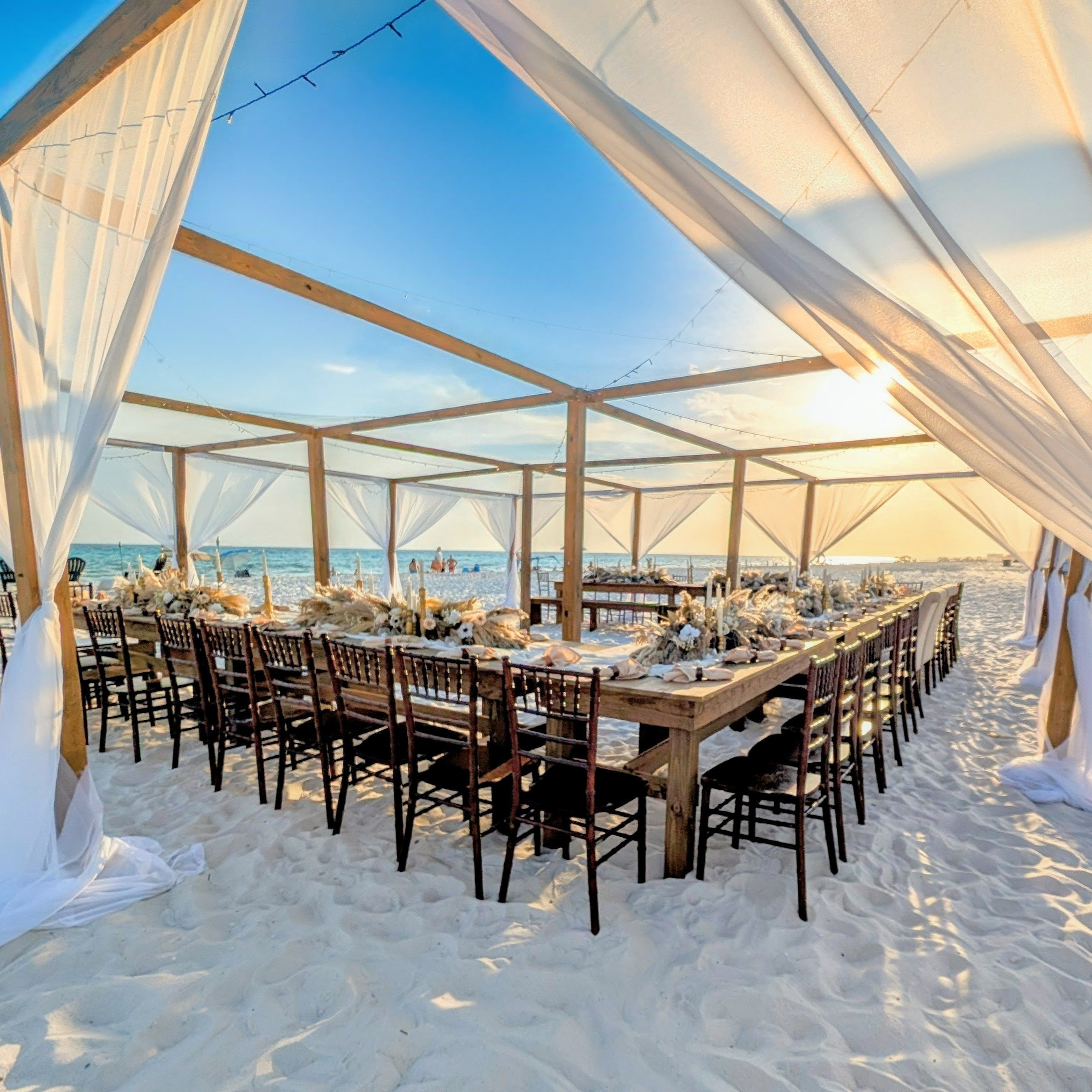 Mahogany chiavari chair rental with farmhouse tables at a beach reception under our exclusive open air tent with beach boho pampas grass centerpiece flowers made by The Wedding Shop