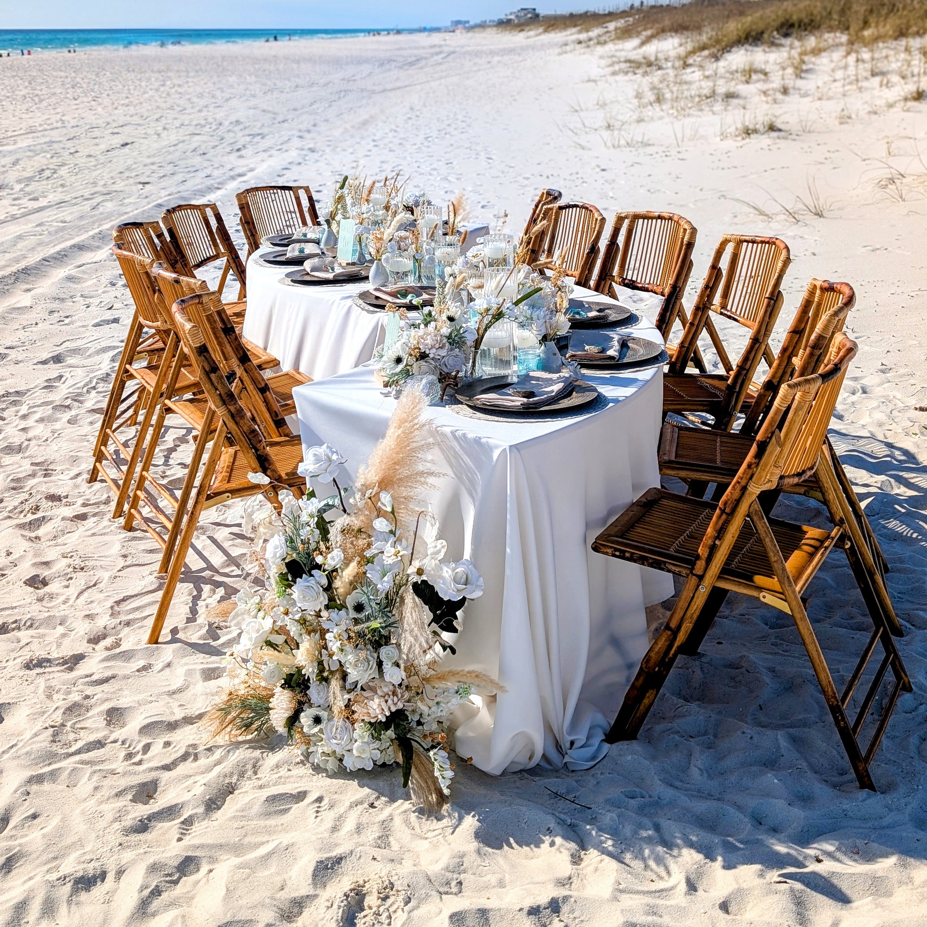 Dining table set for a meal on a sandy beach with chairs and floral decorations and serpentine tables.