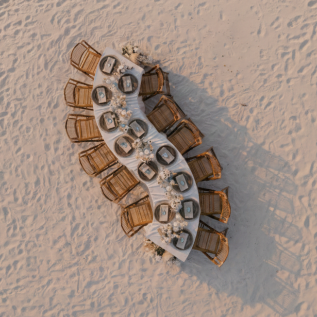 Aerial view of a wedding setup with chairs and serpentine tables on sand.