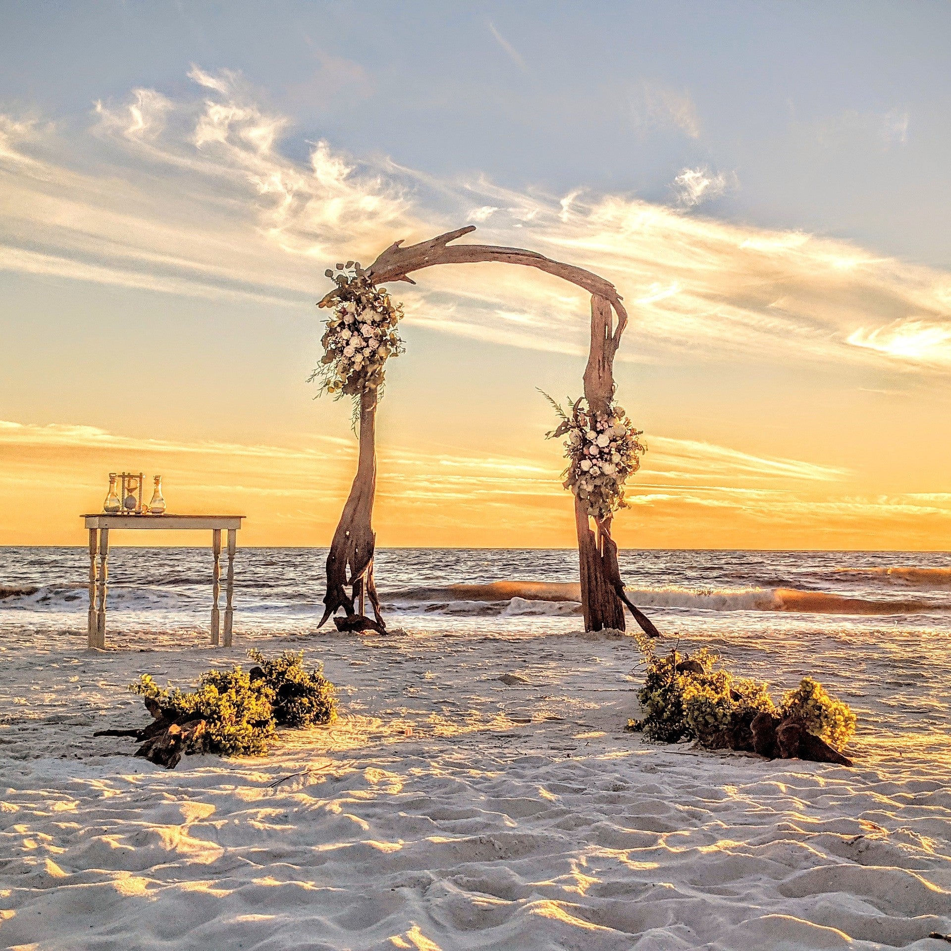 Driftwood arch with floral decorations on a beach at sunset