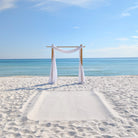 Beach setup with a wooden arch and white fabric on a sandy beach with ocean and sky in the background.