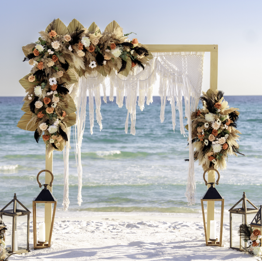 Decorative arch with floral arrangements and lanterns on a beach.