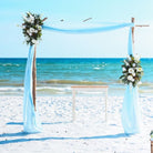 Beach wedding arch with floral decorations and blue drapes against a clear blue sky.