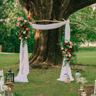 Decorative wood branch wedding arch rental with flowers and white fabric under a large tree in a park setting.