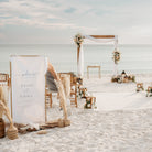 Beach wedding setup with arch, chairs, and decorative elements on a sandy beach.