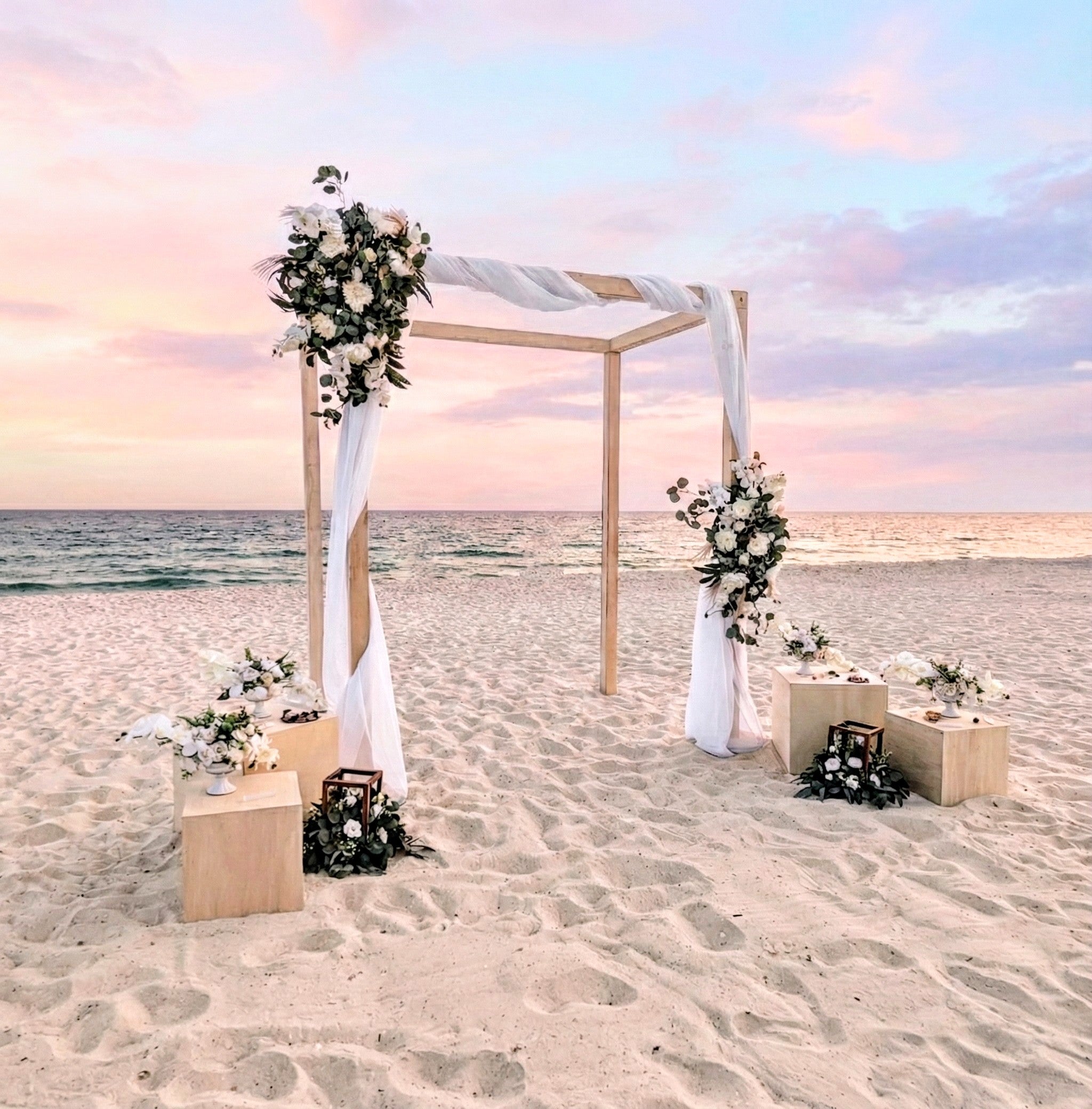 Beach wedding setup with floral arch and decorations on a sandy beach at sunset.