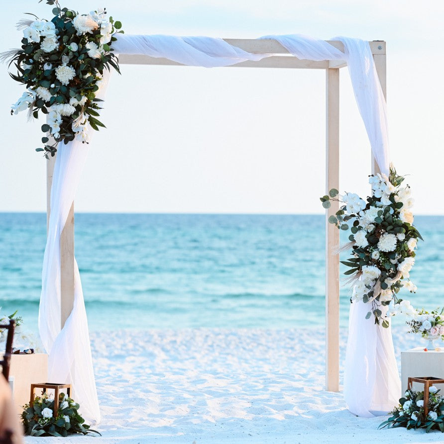 Beach wedding arch decorated with flowers and white drapes, set against a clear blue ocean.