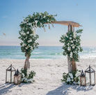 Driftwood wedding arch with flowers and lanterns on a sandy beach with ocean view