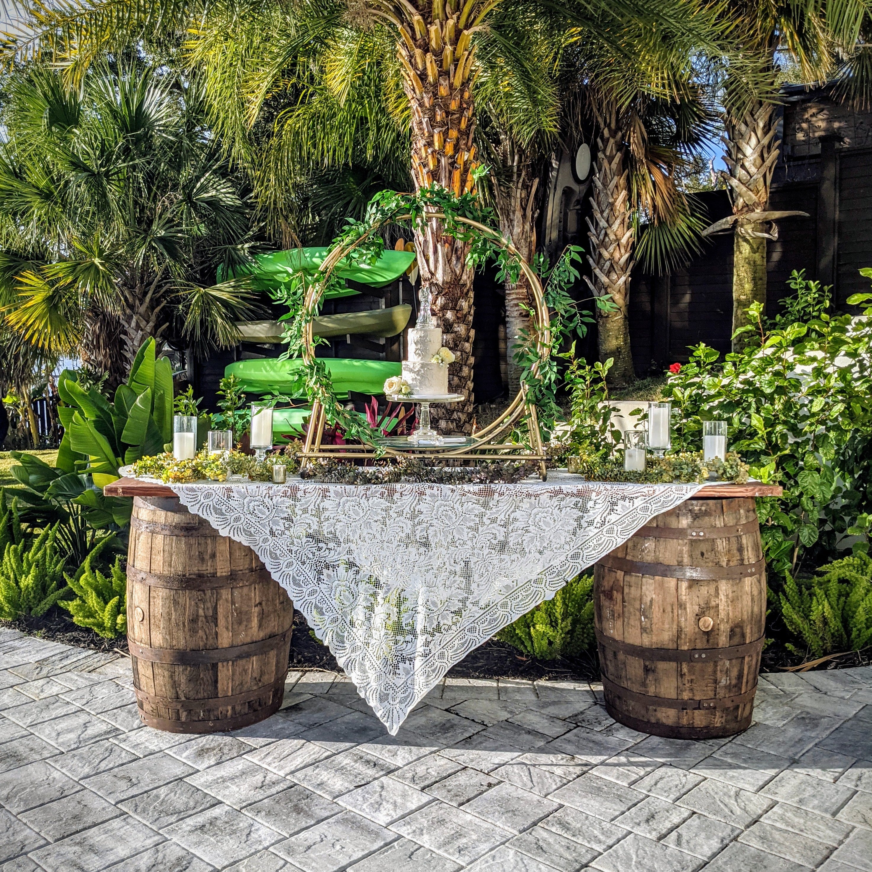 Decorative table rental setup with barrels and lace tablecloth in a garden setting with wedding cake