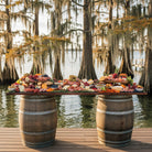 Table with a variety of food displayed on barrels by a body of water with trees.