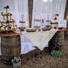 Decorative outdoor table setting with cake stands and a lace tablecloth, surrounded by barrels and plants.