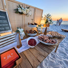 Beachside dining setup with food and drinks on a wooden table, sunset in the background at Inlet Beach in 30A