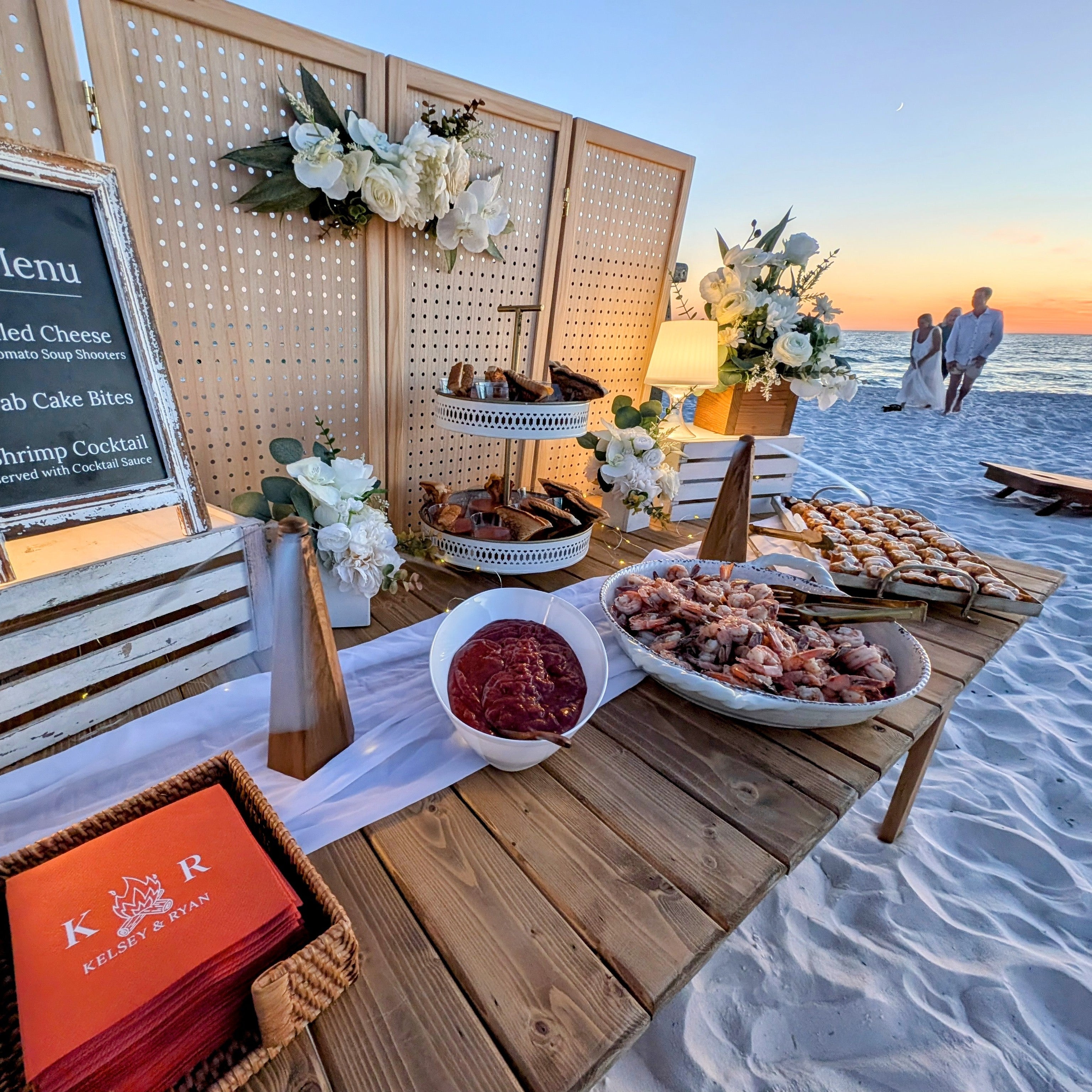 Beachside dining setup with food and drinks on a wooden table, sunset in the background at Inlet Beach in 30A