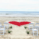 Heart-shaped arrangement of red petals on a sandy beach with white chairs and tables.