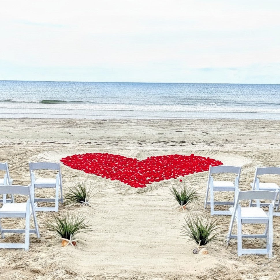 Heart-shaped arrangement of red petals on a sandy beach with white chairs and tables.