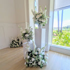 Decorative white wedding flower arrangements on stands in a room with large windows showing greenery outside.