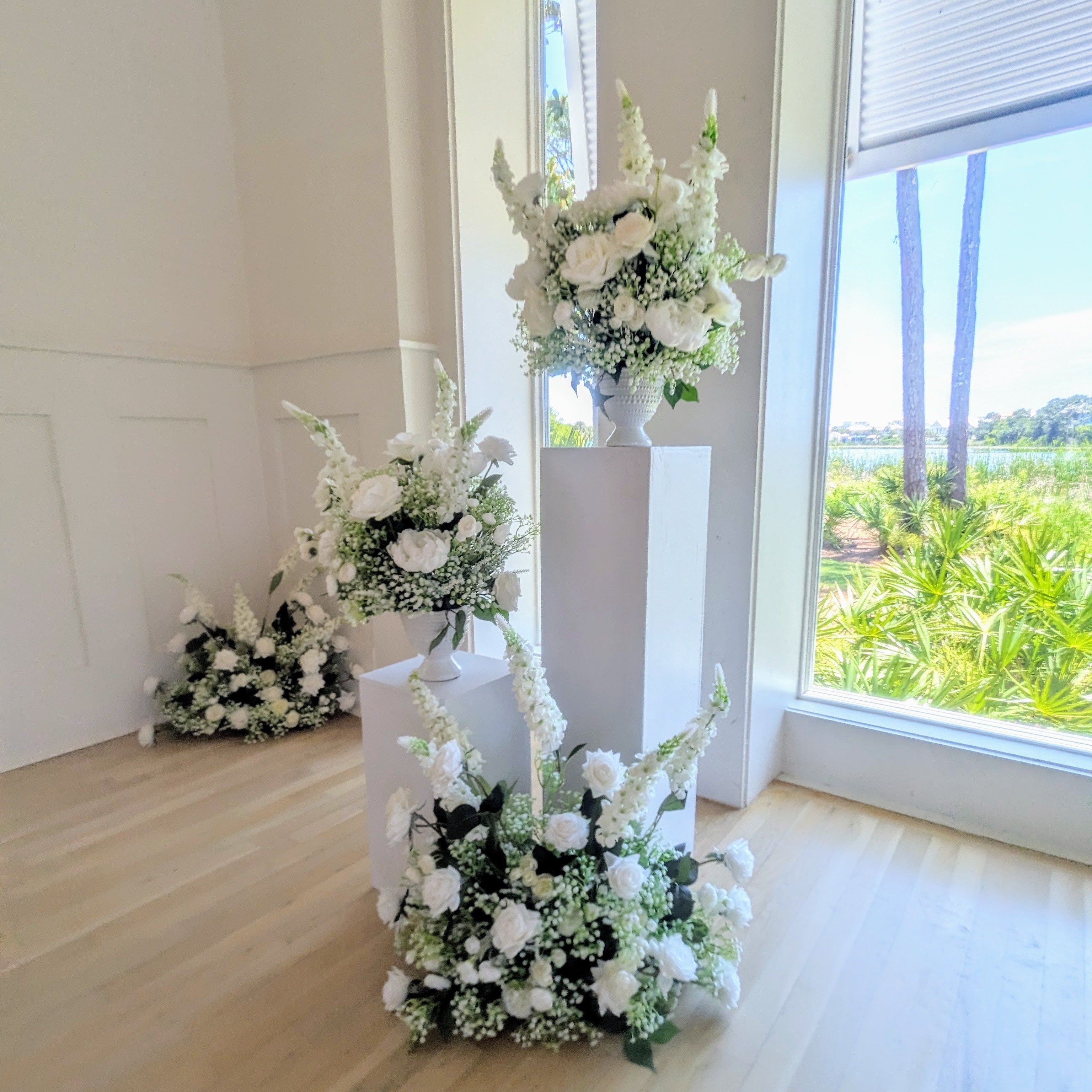 Decorative white wedding flower arrangements on stands in a room with large windows showing greenery outside.