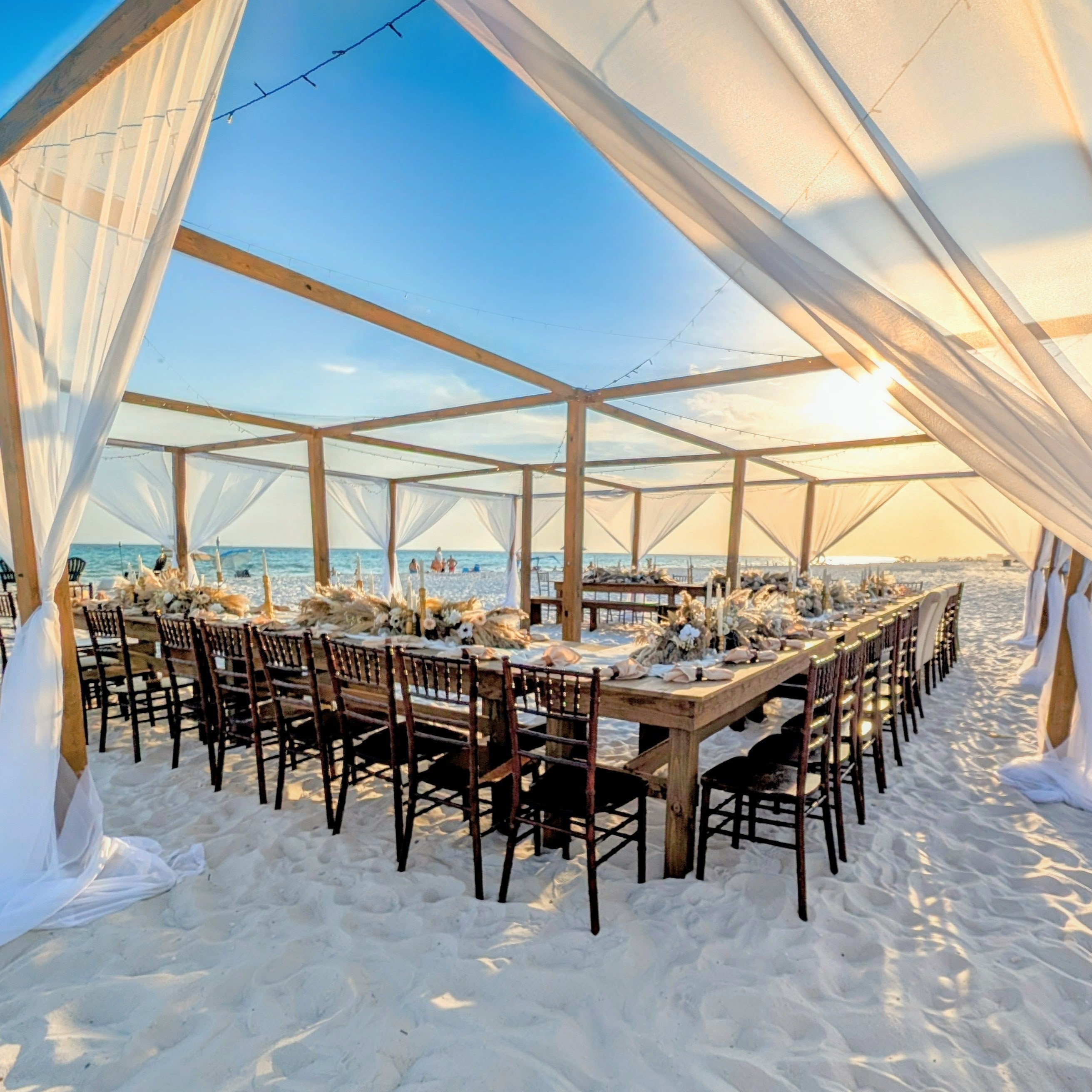 Mahogany chiavari chair rental with farmhouse tables at a beach reception under our exclusive open air tent with beach boho pampas grass centerpiece flowers made by The Wedding Shop