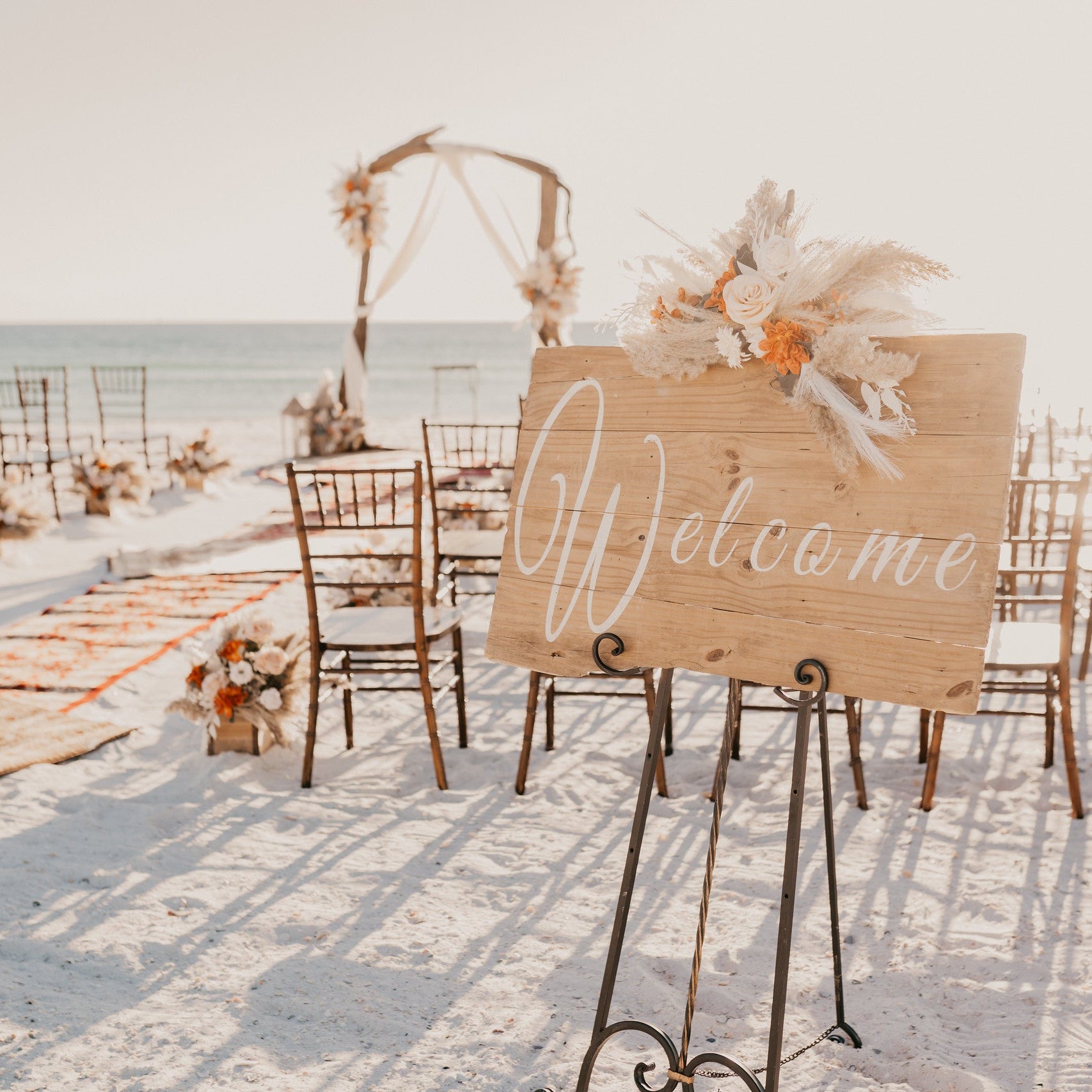 Fall beach wedding ceremony decorations for a destination wedding in Panama City Beach Florida with mahogany chiavari chair rentals, wooden welcome signe, driftwood arbor, aisle runner rugs, and custom wedding flowers by The Wedding Shop