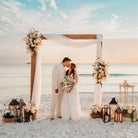 Couple standing under a wedding arch on a beach with floral decorations and lanterns.