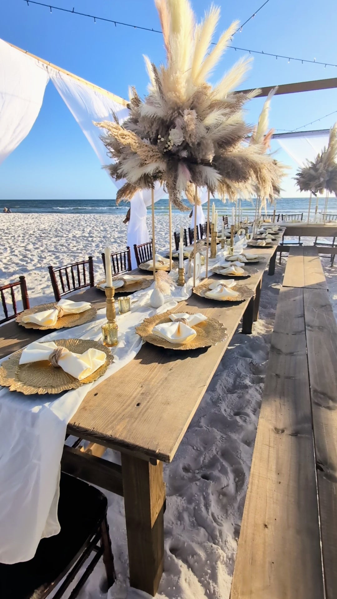 Brown wood bench rentals in Panama City Beach paired with our farmhouse tables for a lux beach reception under our open air tent in PCB with big boho pampas grass centerpieces rented from The Wedding Shop.