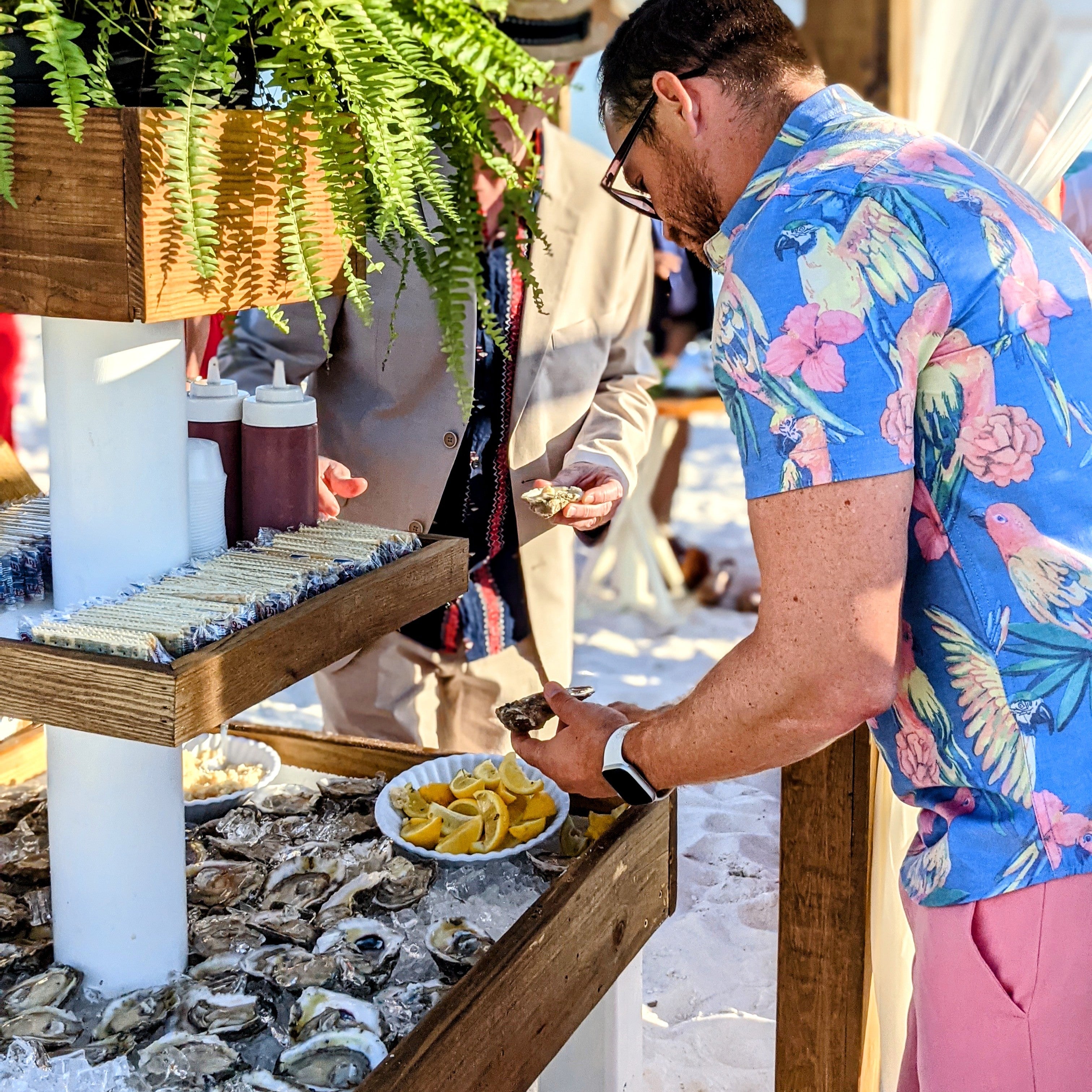 Man in a floral shirt at an outdoor seafood station with oysters and lemon wedges.
