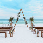 Beach wedding setup with a triangular wooden arch, lanterns, and floral arrangements on a sandy beach.