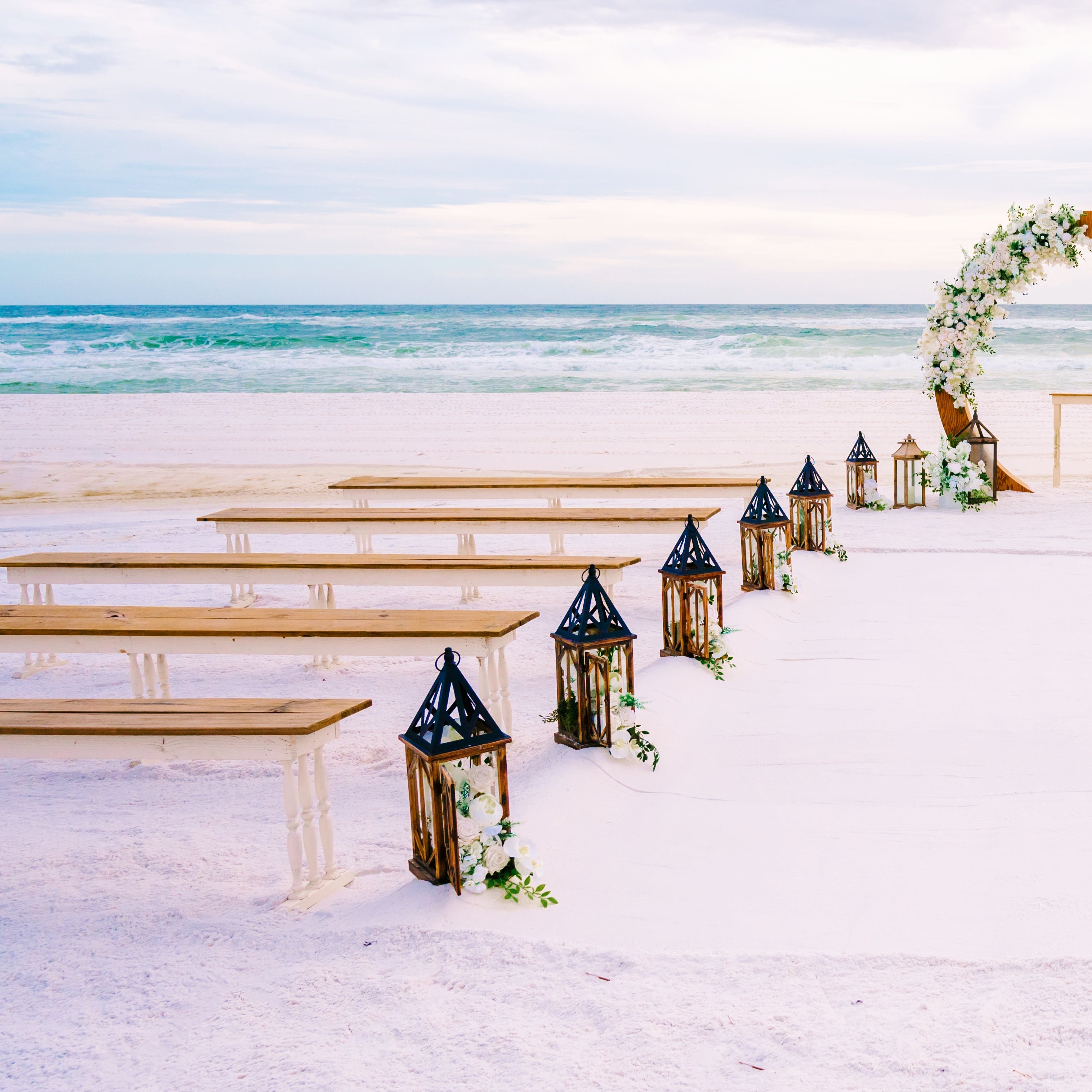 2-toned spindle leg bench seating rental for a beach wedding ceremony with wood circle arbor lantern clusters and white flowers in lanterns for aisle markers by the weddings shop for a beach wedding in panama city beach on the emerald coast