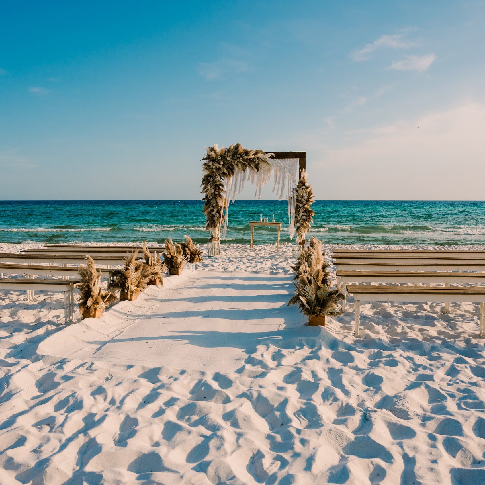 2-toned spindle leg bench seating rental for a beach wedding ceremony with wood arbor and macrame drape and beachy boho flowers by the weddings shop for a beach wedding in panama city beach on the emerald coast