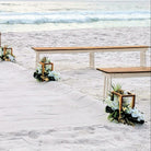 2-toned spindle leg bench seating rental for a beach wedding ceremony with tropical open wood frame aisle marker flowers by the weddings shop for a beach wedding in panama city beach on the emerald coast