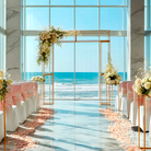 Wedding setup with floral arrangements and chairs by a large window with a view of the ocean.