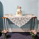Two-tier wedding cake on a table with lace runner, surrounded by floral arrangements and candles against a light blue wall.