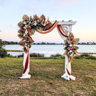 Decorative arch with floral arrangements and red ribbons by a lake at sunset.