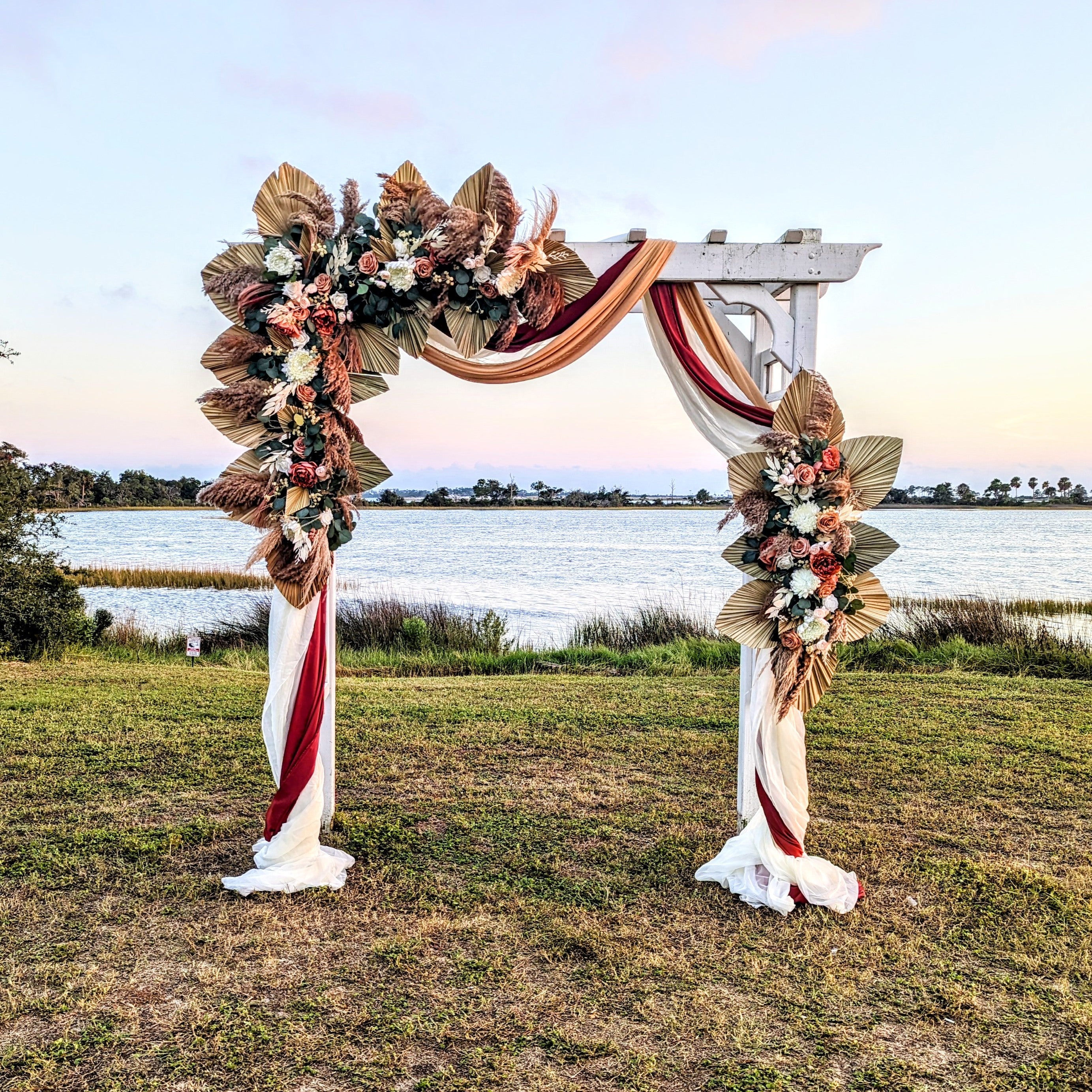 Decorative arch with floral arrangements and red ribbons by a lake at sunset.