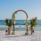 Beach wedding setup with floral arch and palm trees by the ocean.