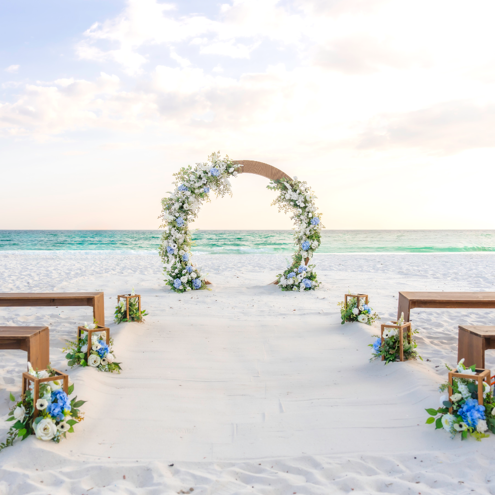 Beach wedding setup with floral arch and chairs under a clear sky.