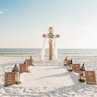 Beach wedding setup with cross, lanterns, and signs on sandy beach.