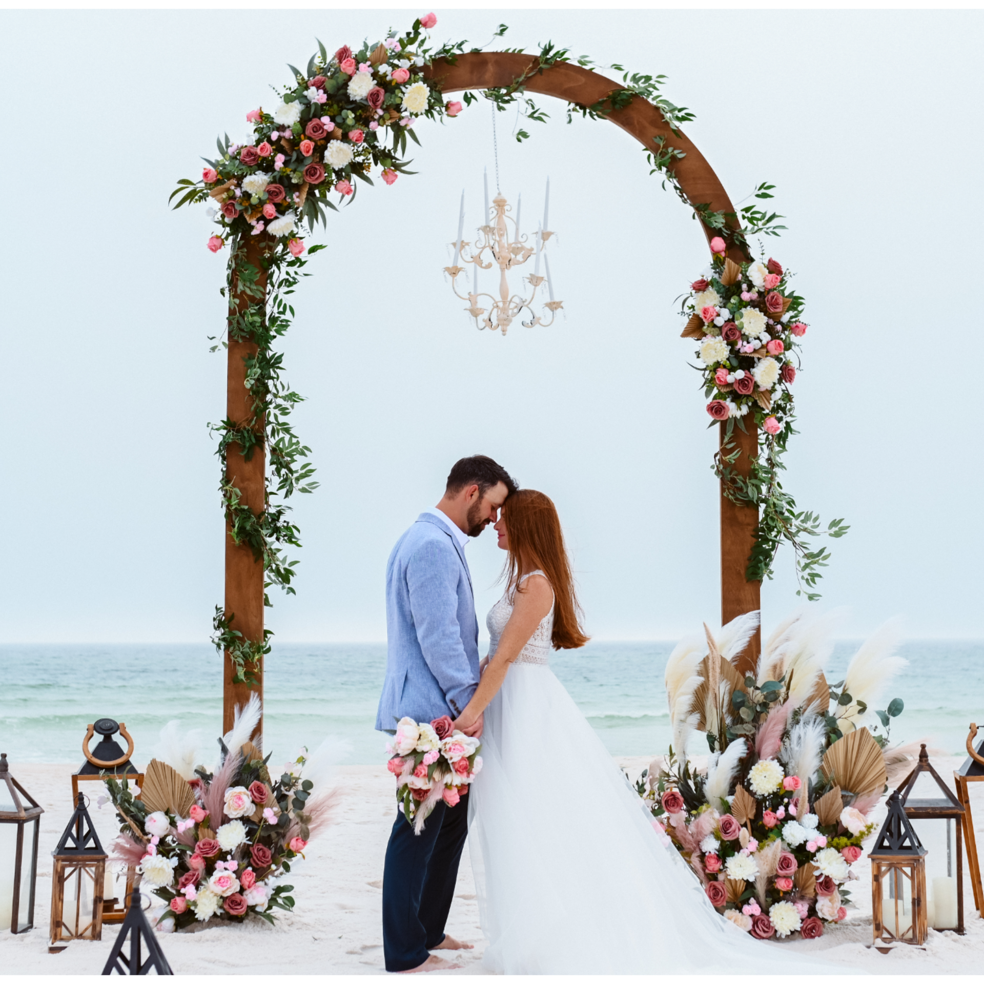 Decorative arch with flowers and a chandelier on a beach, with a couple standing underneath.